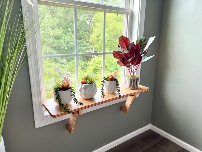 May include: A wooden window shelf with four potted plants. The shelf is mounted on the wall below a window with a view of green trees. The plants are in white ceramic pots, and the shelf is made of light wood.
