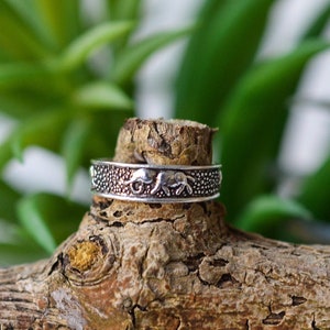 Può includere: Un anello d'argento con una fascia testurizzata e due figure di elefanti. L'anello è appoggiato su un pezzo di legno.