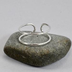 Puede incluir: Un brazalete de plata de doble anillo con un diseño simple y moderno. El brazalete está hecho de dos bandas finas de metal que se unen en los extremos. El brazalete se muestra sobre una roca gris.