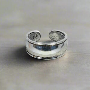 May include: A silver adjustable ring with a wide band and a smooth, polished finish. The ring is marked with "925" on the inside.