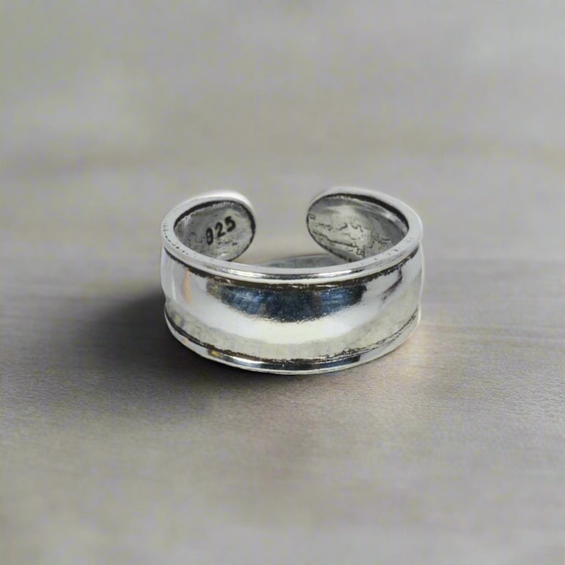 May include: A silver adjustable ring with a wide band and a smooth, polished finish. The ring is marked with "925" on the inside.