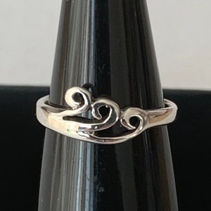 Puede incluir: Un anillo de plata con un diseño calado en espiral. El anillo presenta un patrón continuo y fluido de curvas y bucles, creando una estética elegante y delicada. El anillo se exhibe sobre un soporte negro sobre un fondo blanco.