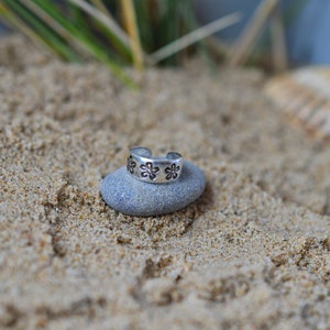 May include: A silver toe ring with a floral design. The ring is sitting on a gray rock.