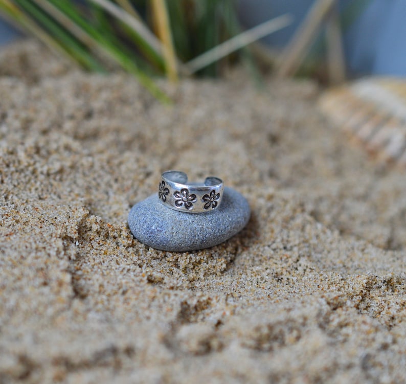 May include: A silver toe ring with a floral design. The ring is sitting on a gray rock.