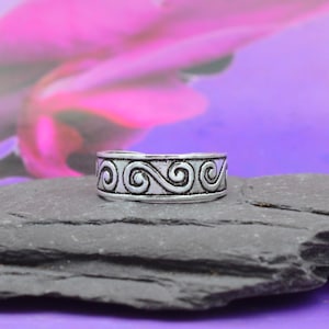 Pode incluir: Um anel prateado com um design de rolagem repetido. O anel está colocado sobre uma pedra cinza escura contra um fundo desfocado de tons rosa e roxo. O design do anel apresenta uma série de padrões de redemoinho interconectados.