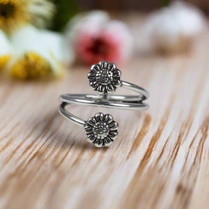 Könnte beinhalten: Ein silberner Ring mit zwei Sonnenblumen-Designs. Der Ring ist verdreht und hat einen zarten, femininen Look.