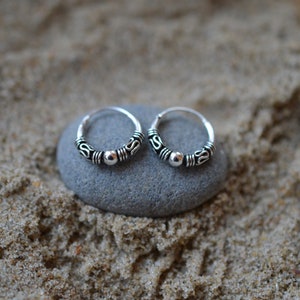 May include: A pair of small silver hoop earrings with a beaded design. The earrings are on a gray stone surface.