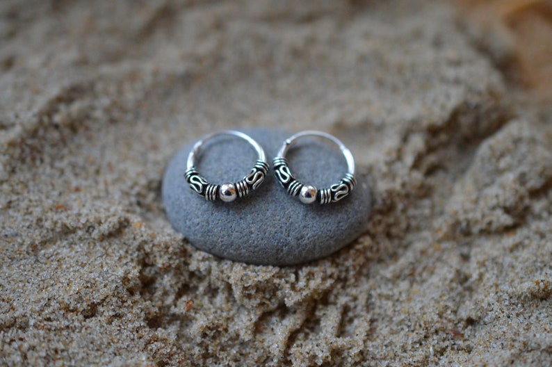 May include: A pair of small silver hoop earrings with a beaded design. The earrings are on a gray stone surface.