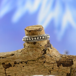 Peut inclure: Une bague en argent avec un motif de perles. La bague présente une bande de petites perles espacées régulièrement entre deux rangées de perles plus grosses. La bague est présentée sur un morceau de bois vieilli sur fond bleu flou.