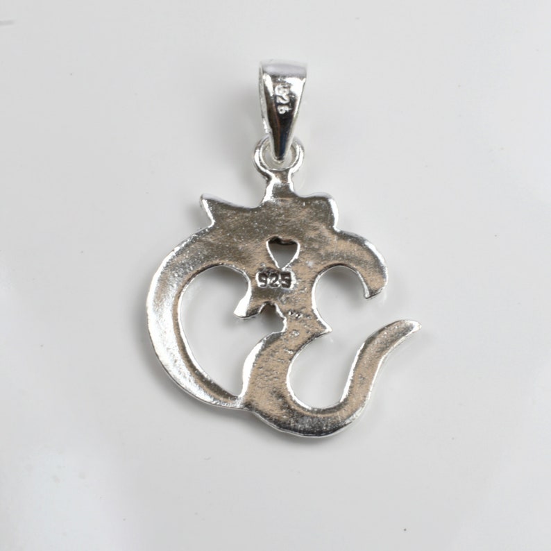 May include: A silver pendant in the shape of the Om symbol, with a loop at the top for a chain. The pendant has a heart-shaped cutout and the marking "925". The pendant is on a white background.