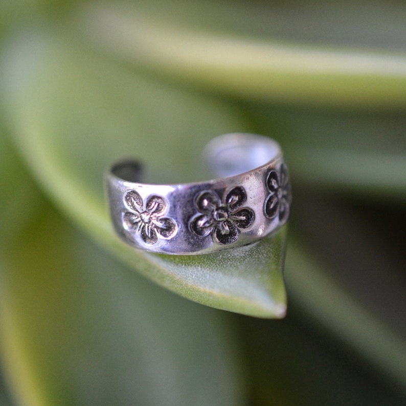 May include: A silver ear cuff with a floral design. The cuff is open and has a rounded shape. It is sitting on a green leaf.