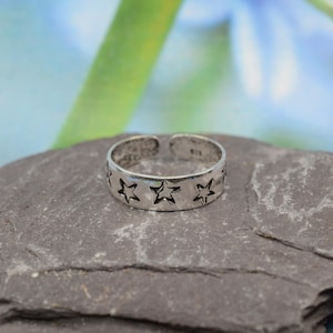 K&ouml;nnte beinhalten: Ein verstellbarer Ring in Silber mit einem Sternmuster. Der Ring hat ausgeschnittene Sternformen um das Band. Der Ring wird auf einer grauen Steinoberfl&auml;che mit einem verschwommenen gr&uuml;n-blauen Hintergrund pr&auml;sentiert.