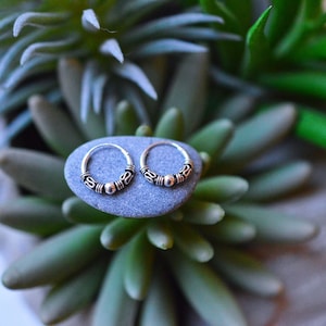 May include: Two silver hoop earrings with a beaded design. The earrings are on a gray stone.