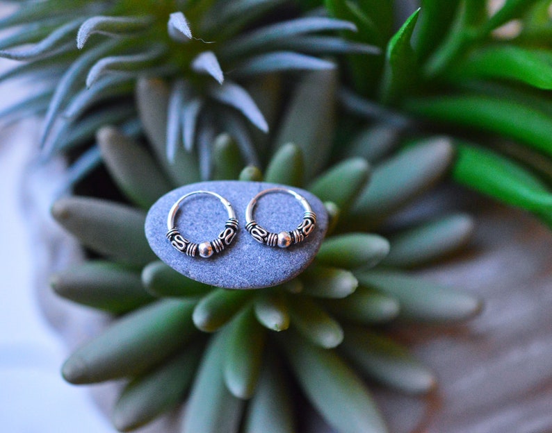 May include: Two silver hoop earrings with a beaded design. The earrings are on a gray stone.