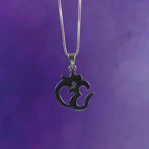 May include: A silver-colored pendant necklace featuring the Om symbol. The pendant is intricately designed with a stylized script and hangs from a delicate chain. The background is a gradient of purple hues.