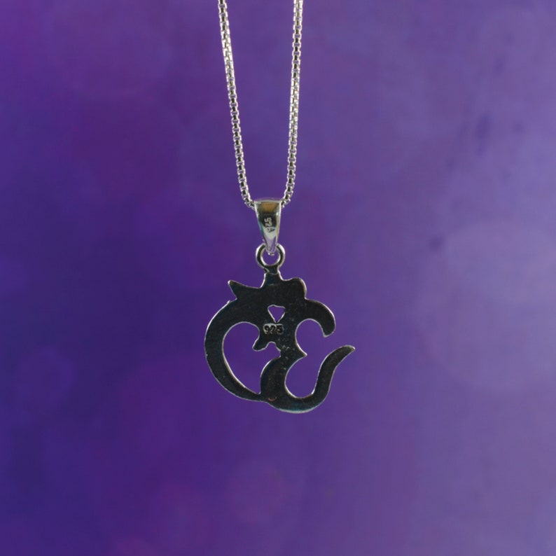 May include: A silver-colored pendant necklace featuring the Om symbol. The pendant is intricately designed with a stylized script and hangs from a delicate chain. The background is a gradient of purple hues.