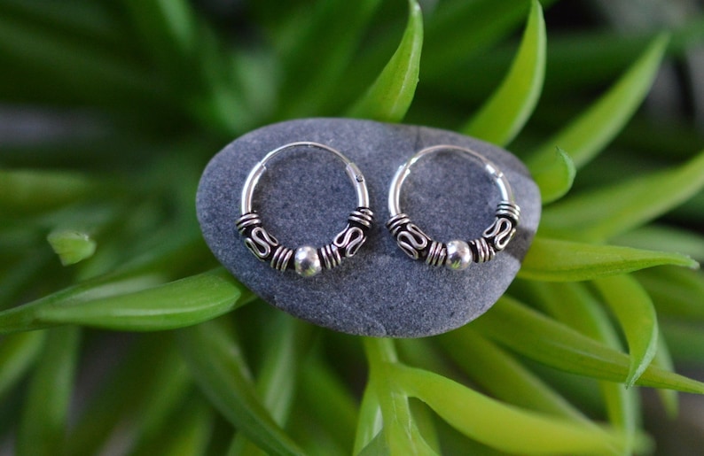 May include: A pair of small silver hoop earrings with a beaded design. The earrings are on a gray stone.