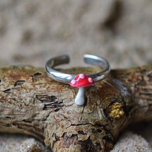 Op de afbeelding: Zilveren verstelbare ring met een kleine, gedetailleerde paddenstoelbedel. De paddenstoel heeft een rode hoed met witte stippen en een witte steel. De ring wordt gepresenteerd op een stuk drijfhout.