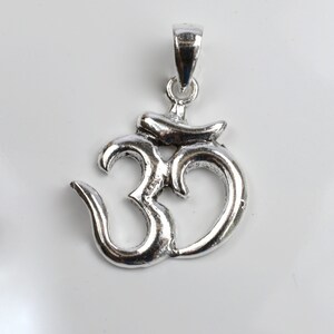 May include: A silver-colored pendant in the shape of the Om symbol. The pendant has a loop at the top for a chain and is displayed against a white background. The Om symbol is a sacred sound and spiritual icon in Hinduism.