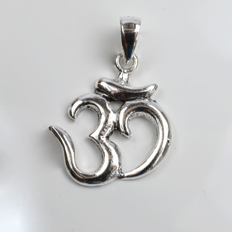 May include: A silver-colored pendant in the shape of the Om symbol. The pendant has a loop at the top for a chain and is displayed against a white background. The Om symbol is a sacred sound and spiritual icon in Hinduism.