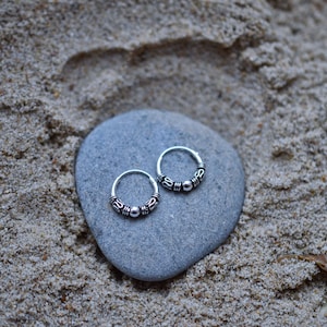 May include: Two silver hoop earrings with a beaded design. The earrings are on a gray stone.