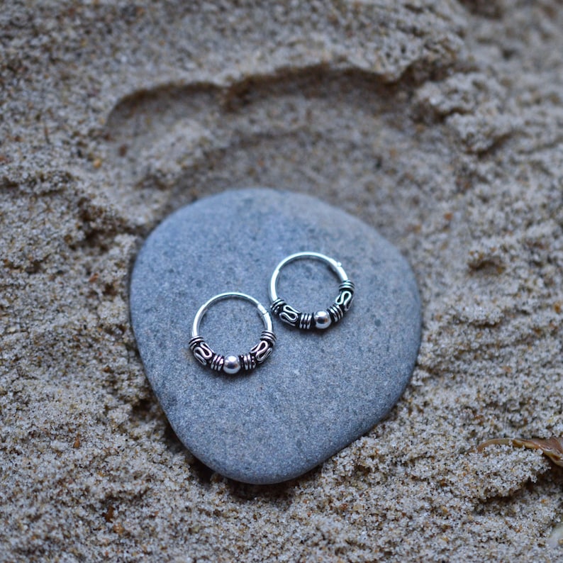 May include: Two silver hoop earrings with a beaded design. The earrings are on a gray stone.