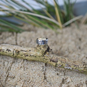 May include: A silver toe ring with a floral design. The ring is sitting on a piece of driftwood.
