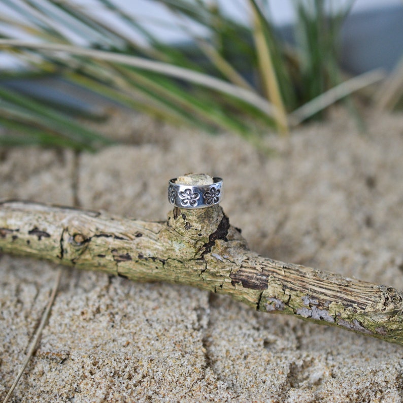 May include: A silver toe ring with a floral design. The ring is sitting on a piece of driftwood.