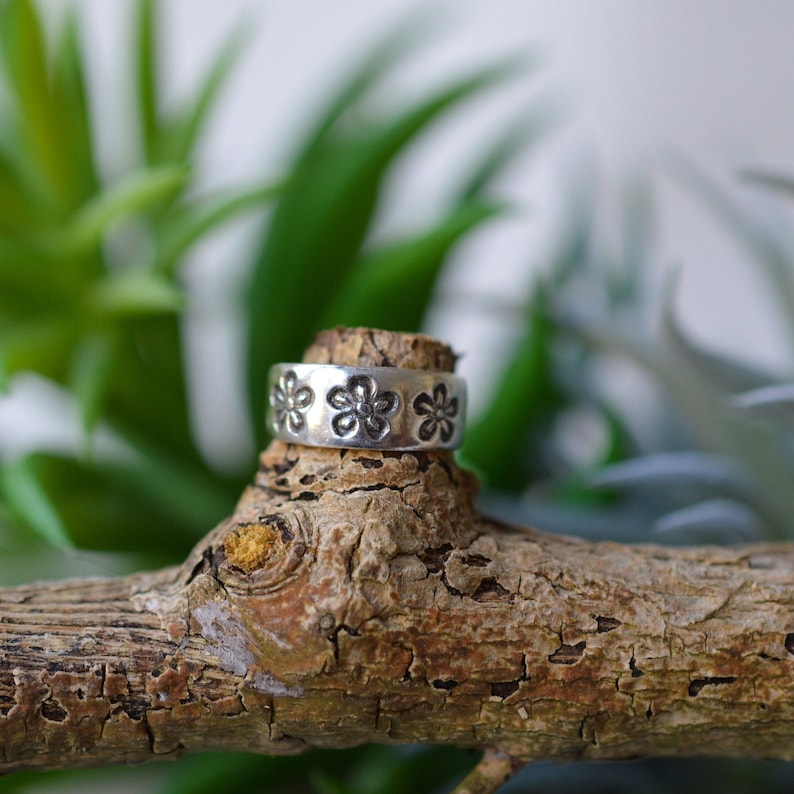 May include: A silver ring with a floral design. The ring is sitting on a piece of wood.