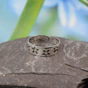 K&ouml;nnte beinhalten: Ein silberfarbener Ring mit Sternmuster. Der verstellbare Ring hat ausgeschnittene Sternformen um das Band. Der Ring wird auf einer grauen Steinoberfl&auml;che mit verschwommenem gr&uuml;n-blauem Hintergrund gezeigt.