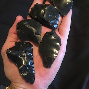 May include: Five pieces of black obsidian, a type of volcanic glass, are shown in a hand. The pieces are irregularly shaped and have a shiny, glassy surface.