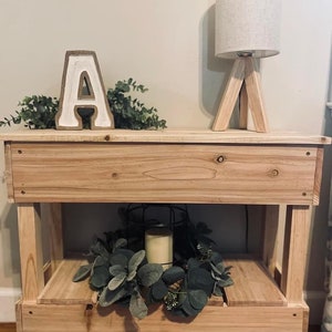 May include: A wooden two-tiered side table with a white letter A, a small lamp with a white linen shade, and a green leafy wreath on the lower shelf.