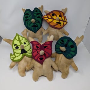 May include: Five plush toys with different coloured masks. The masks are all different shapes and have different facial expressions. The toys are all standing on a white surface.