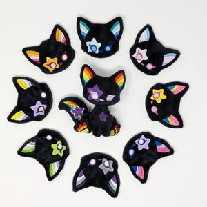 Puede incluir: Colección de peluches y parches negros con forma de gato y detalles coloridos. Las figuras de gato presentan orejas, colas y diseños en forma de estrella con colores arcoíris. Dispuestos en círculo sobre un fondo blanco.
