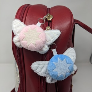 May include: Two plush keychains with white wings and a star in the center. The keychain on the left has a pink star and the keychain on the right has a blue star. The keychains are attached to a red backpack.