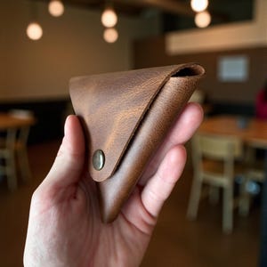 May include: A brown leather wallet with a triangular shape and a snap closure. The wallet has a textured surface and appears to be made of high-quality leather. The wallet is designed to hold cards and cash.