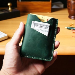 Peut inclure: Un porte-cartes en cuir vert foncé avec une bordure cousue vert clair. Le porte-cartes contient une carte de crédit et un billet de cent dollars américains. Le porte-cartes est tenu dans la main d'une personne.