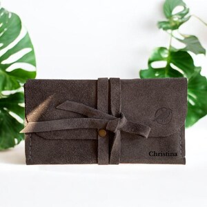 Handmade green suede leather wallet with soft texture and snap button closure, shown being held by hand. The wallet has a minimalist, long clutch design ideal for carrying cards, cash, or small essentials. Styled with a clean white wood background and green succulents, highlighting the natural and elegant feel of the product. A perfect choice for daily use or casual minimalist fashion lovers.