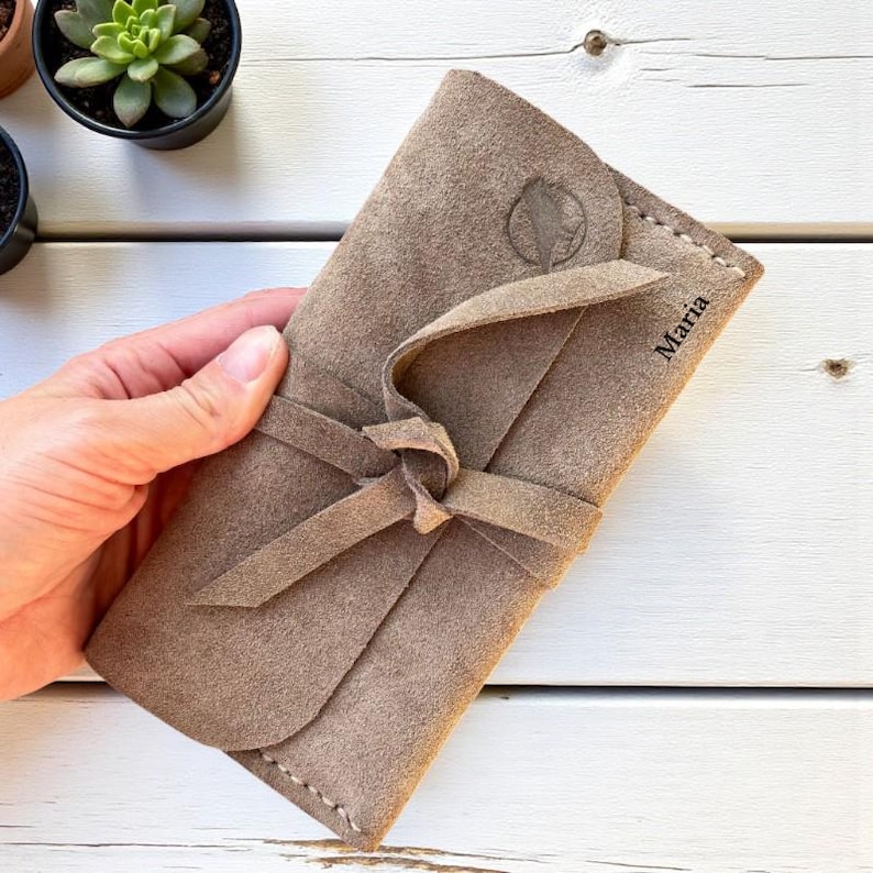 Handmade green suede leather wallet with soft texture and snap button closure, shown being held by hand. The wallet has a minimalist, long clutch design ideal for carrying cards, cash, or small essentials. Styled with a clean white wood background and green succulents, highlighting the natural and elegant feel of the product. A perfect choice for daily use or casual minimalist fashion lovers.