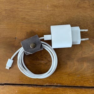 May include: A white USB-C charging cable and a white power adapter are displayed. A small, dark gray leather cable organizer with a brass snap is also present. The items are arranged on a wooden surface.