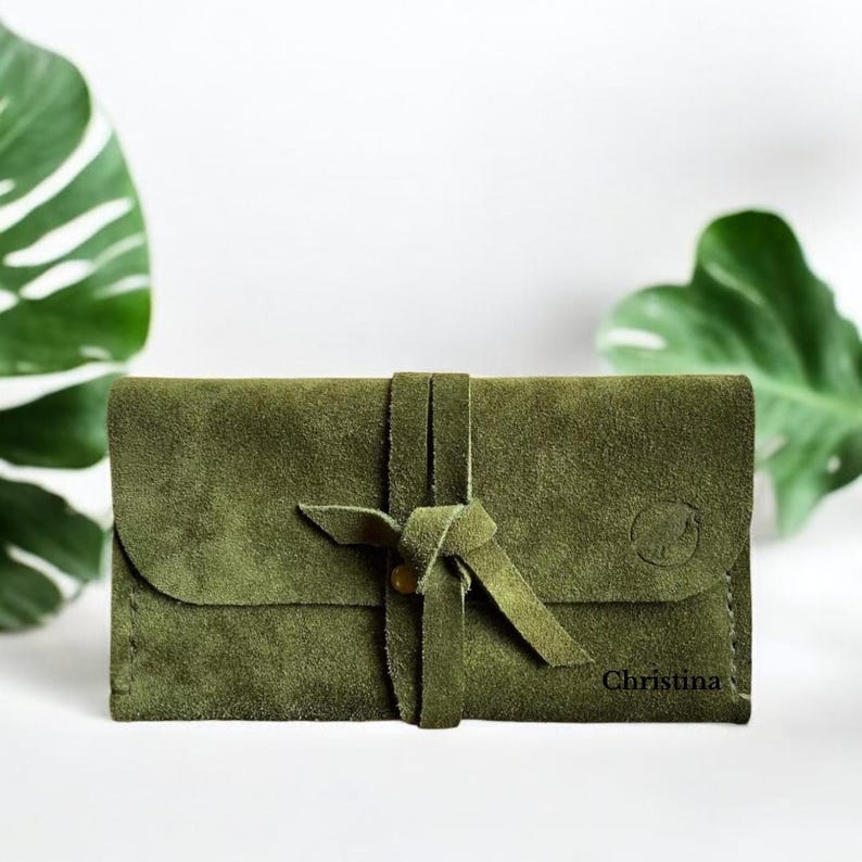 Handmade green suede leather wallet with soft texture and snap button closure, shown being held by hand. The wallet has a minimalist, long clutch design ideal for carrying cards, cash, or small essentials. Styled with a clean white wood background and green succulents, highlighting the natural and elegant feel of the product. A perfect choice for daily use or casual minimalist fashion lovers.