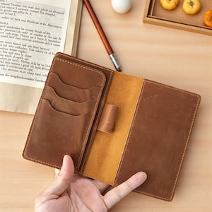 以下が含まれることがあります： 複数のカードスロットとペンホルダーが付いた、開いた茶色の革製ジャーナル。ジャーナルは開いており、明るい茶色の内側が見えます。木製のペンが表面に置かれています。ジャーナルは手で持たれています。