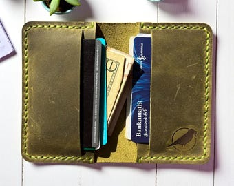 May include: Open olive green leather wallet with a bird logo, holding cards and cash. The wallet has a stitched border and is open, revealing the interior. The background is a white wooden surface with small potted plants.