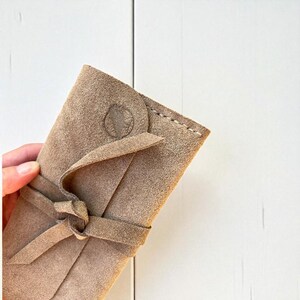 Handmade green suede leather wallet with soft texture and snap button closure, shown being held by hand. The wallet has a minimalist, long clutch design ideal for carrying cards, cash, or small essentials. Styled with a clean white wood background and green succulents, highlighting the natural and elegant feel of the product. A perfect choice for daily use or casual minimalist fashion lovers.