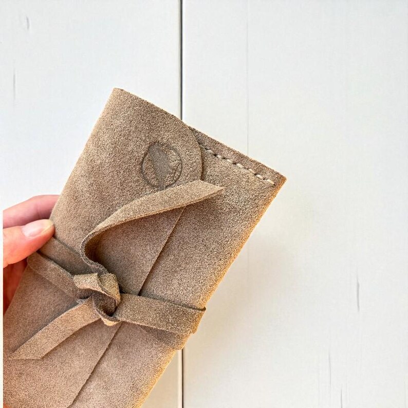 Handmade green suede leather wallet with soft texture and snap button closure, shown being held by hand. The wallet has a minimalist, long clutch design ideal for carrying cards, cash, or small essentials. Styled with a clean white wood background and green succulents, highlighting the natural and elegant feel of the product. A perfect choice for daily use or casual minimalist fashion lovers.