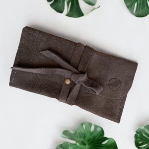 Handmade green suede leather wallet with soft texture and snap button closure, shown being held by hand. The wallet has a minimalist, long clutch design ideal for carrying cards, cash, or small essentials. Styled with a clean white wood background and green succulents, highlighting the natural and elegant feel of the product. A perfect choice for daily use or casual minimalist fashion lovers.