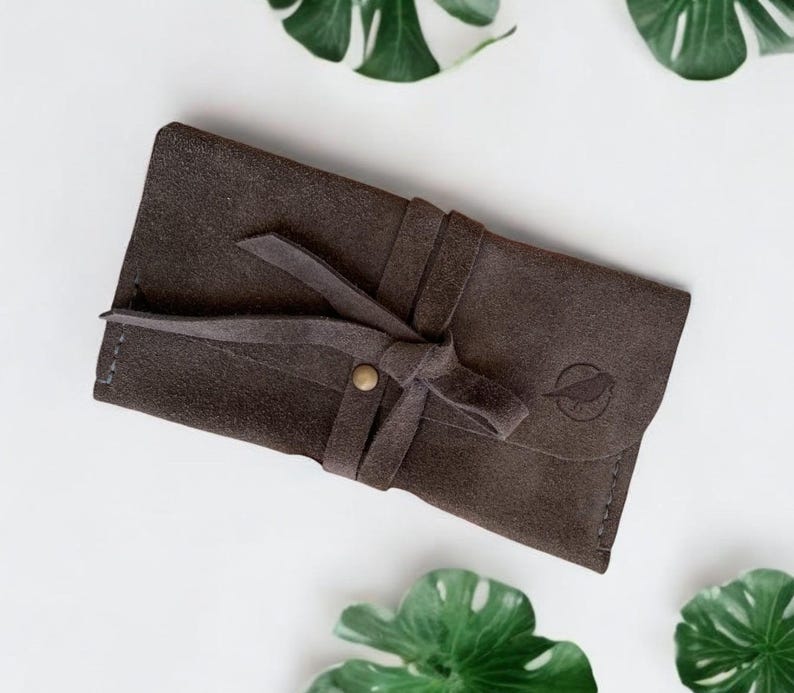 Handmade green suede leather wallet with soft texture and snap button closure, shown being held by hand. The wallet has a minimalist, long clutch design ideal for carrying cards, cash, or small essentials. Styled with a clean white wood background and green succulents, highlighting the natural and elegant feel of the product. A perfect choice for daily use or casual minimalist fashion lovers.