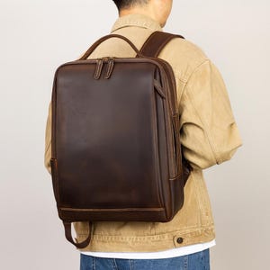 May include: A brown leather backpack with a rectangular shape and a zipper closure. The backpack has two shoulder straps and a top handle. The leather has a distressed look.