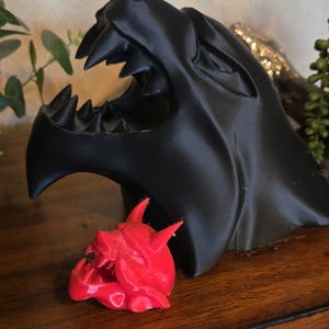 May include: A large matte black 3D printed sculpture of a snarling wolf head with an open mouth showing sharp teeth, positioned behind a smaller, bright red 3D printed sculpture of a horned demon head with an open mouth and visible fangs.