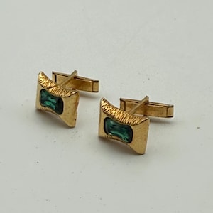 May include: A pair of gold-toned cufflinks featuring a rectangular, textured metal frame surrounding a central, emerald-green rectangular stone. Each cufflink has a hinged T-bar toggle mechanism attached to the back for securing through a shirt cuff.